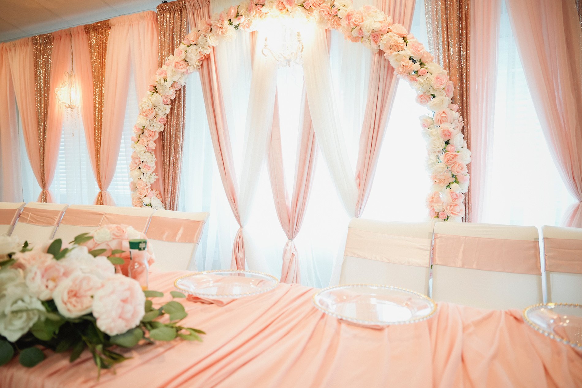 Elegant pink and white wedding backdrop with floral arch, draped curtains, and chandelier, set for reception or ceremony.