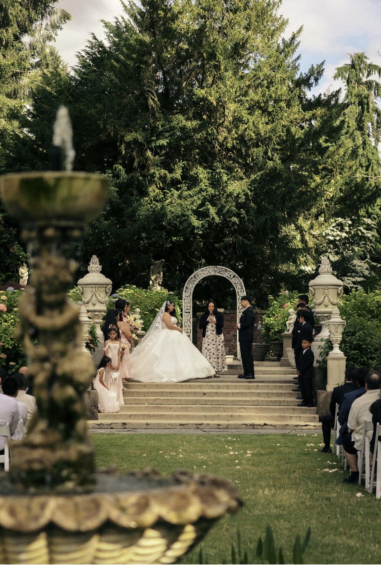 A wedding ceremony outdoors in a garden with green trees, stone steps, and ornate white arches. The bride in a white gown and veil is standing with the groom in a dark suit. Several children and adults are present, some sitting and some standing, wit
