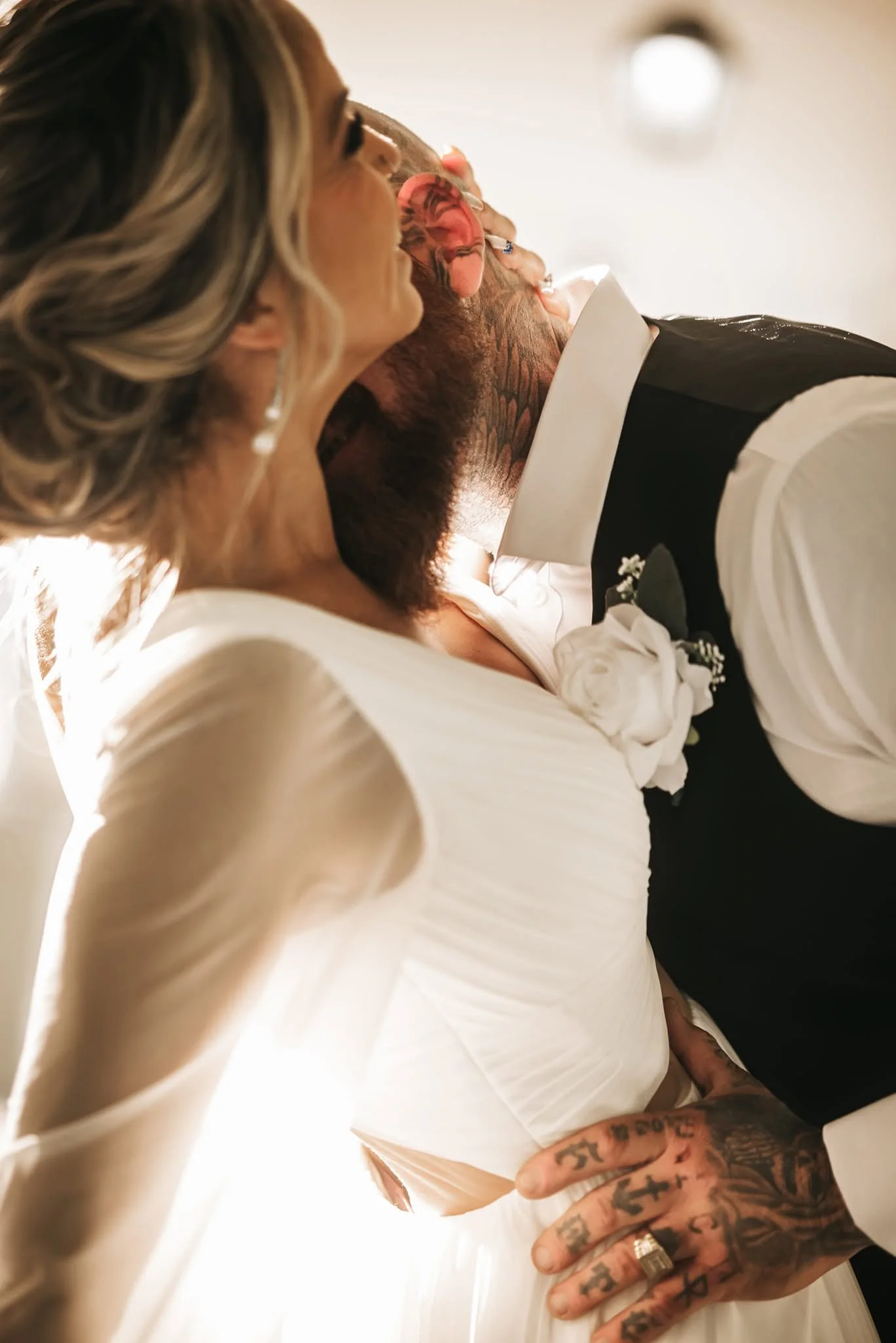Close-up of a person with blonde hair, tattoos on their hand and neck, wearing rings, with another person in black and white clothing leaning on their shoulder. The background is blurred with soft lighting.