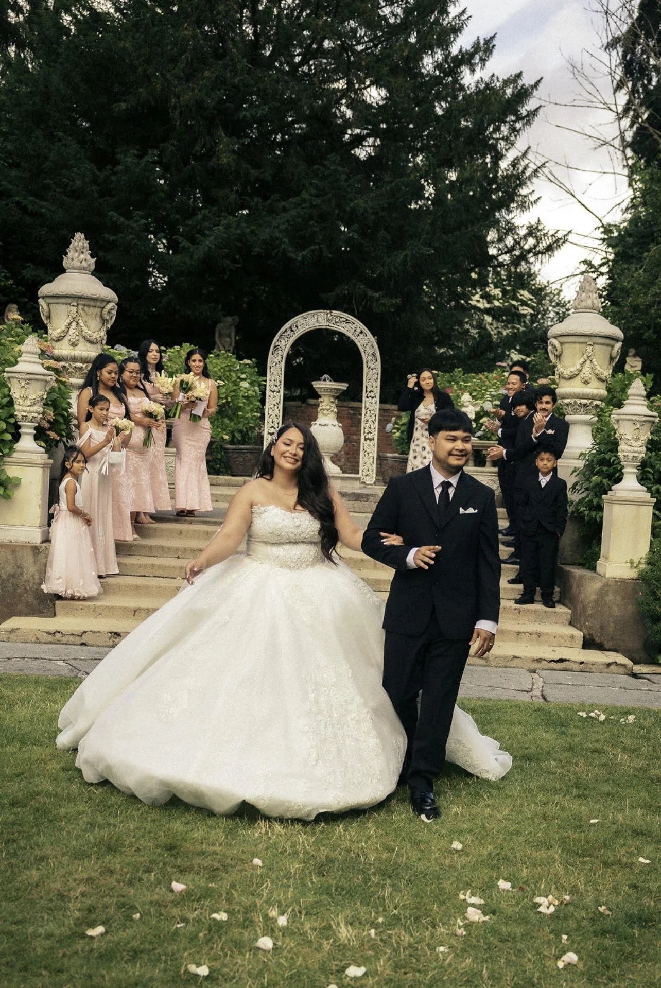 A bride in a white wedding gown and a groom in a black tuxedo walking on grass, with wedding guests on stairs and in the background, some holding flowers, at an outdoor wedding ceremony.