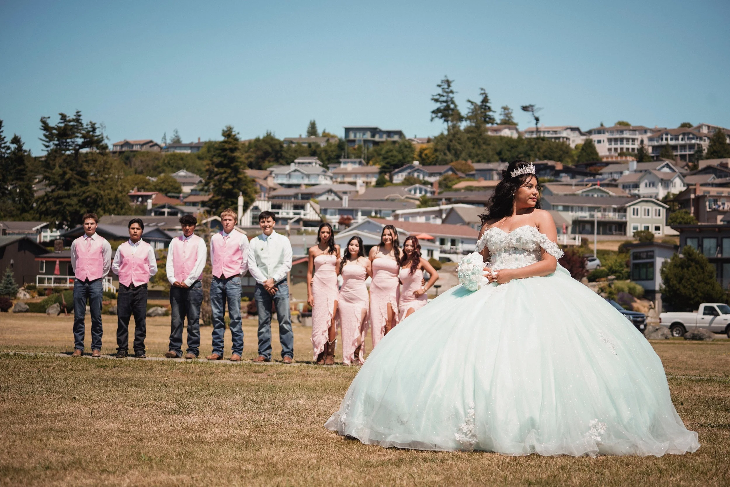 A bride in a white wedding gown with a tiara stands in front of bridesmaids and groomsmen during an outdoor wedding ceremony on a grassy field with houses on a hillside in the background.