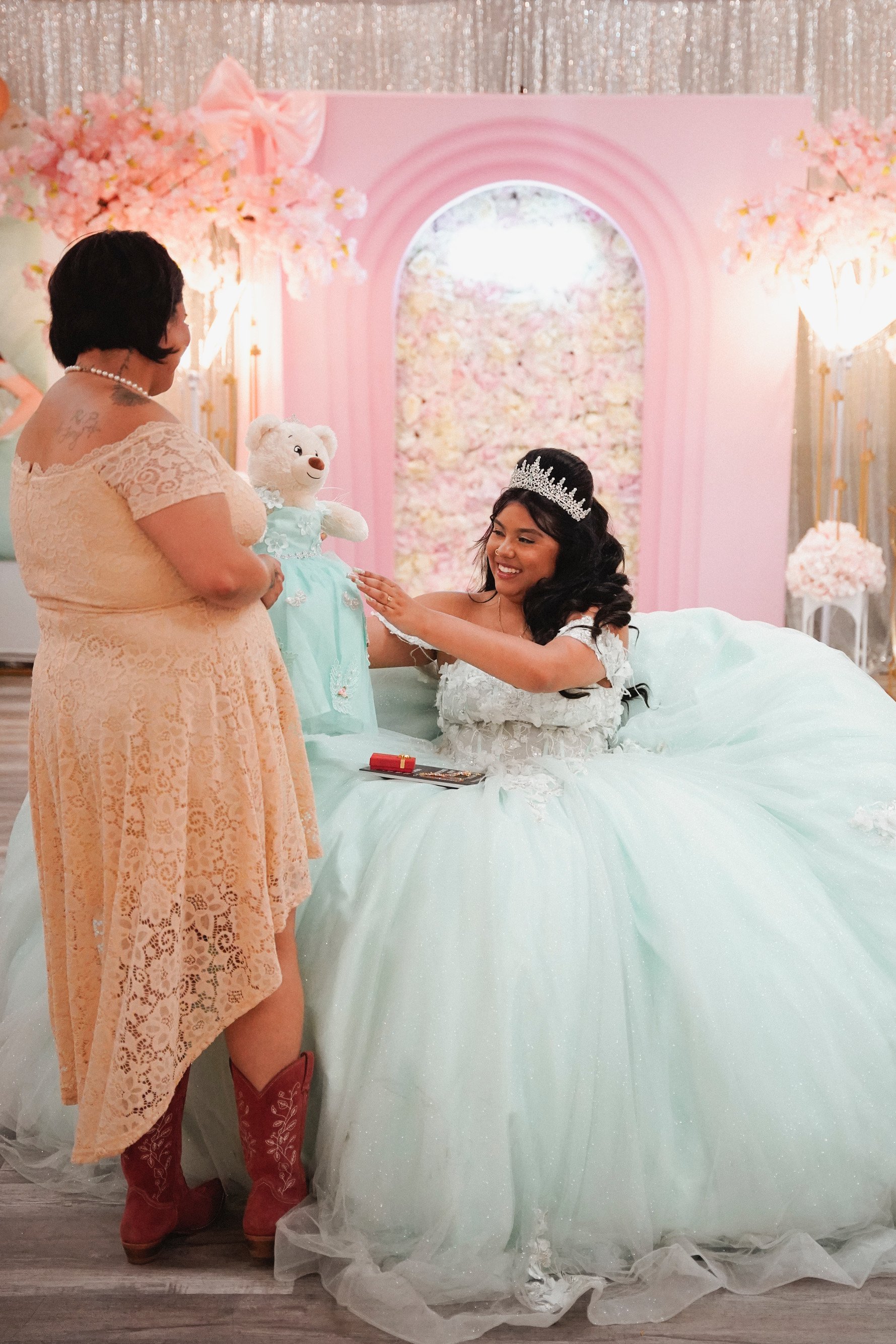 A young woman in a white dress and tiara sitting on a pink and white bed, smiling as she touches a white teddy bear dressed in a light blue outfit. A woman in a peach lace dress with red cowboy boots stands next to her, holding the teddy bear. The ba