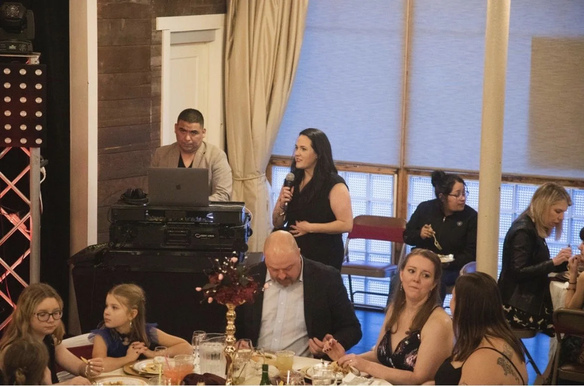 People at a social gathering with a woman speaking into a microphone, others sitting at tables, and a DJ setup in the background.