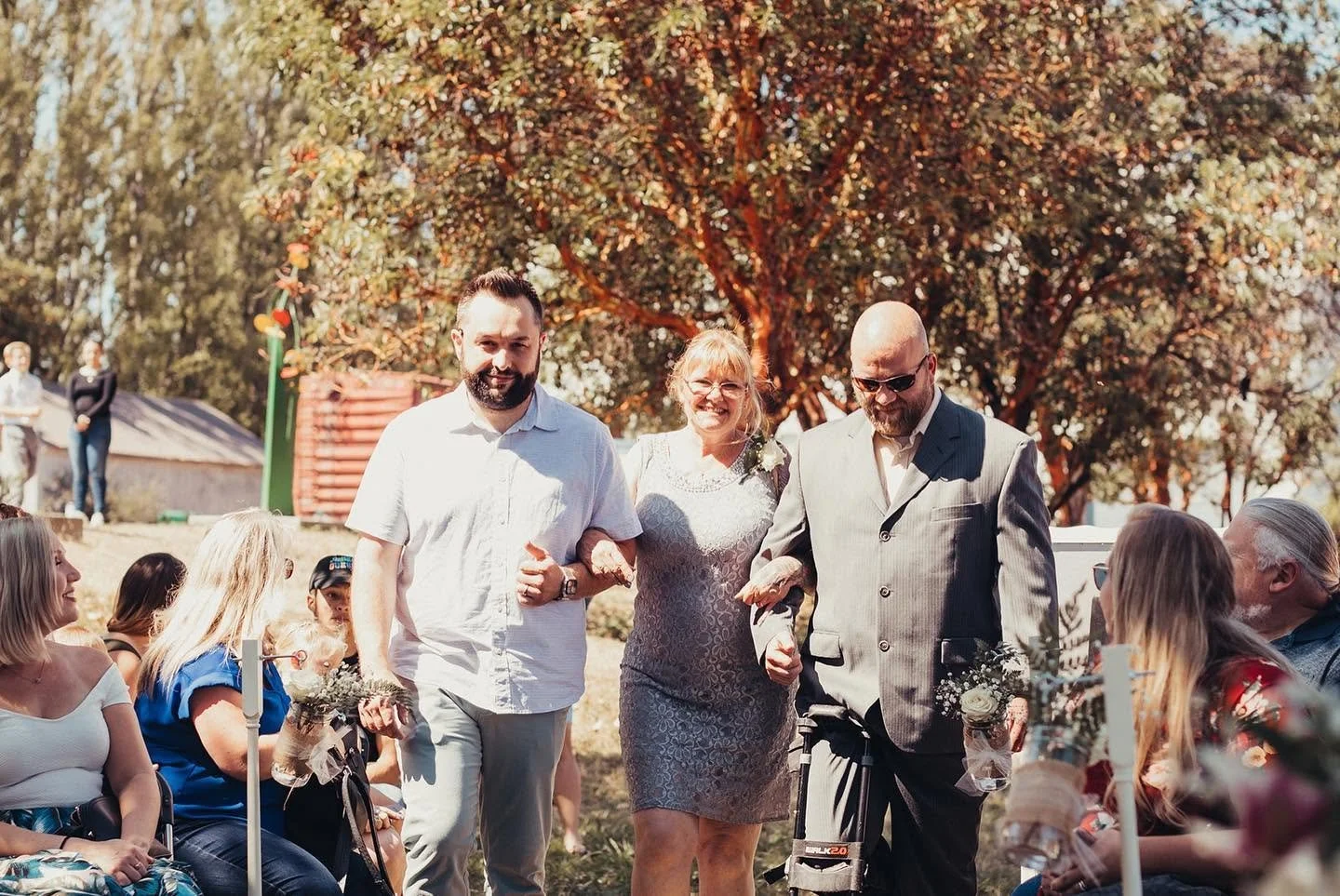 Three people walking arm-in-arm at an outdoor event, with guests seated around them, in front of a large tree with orange and green foliage.