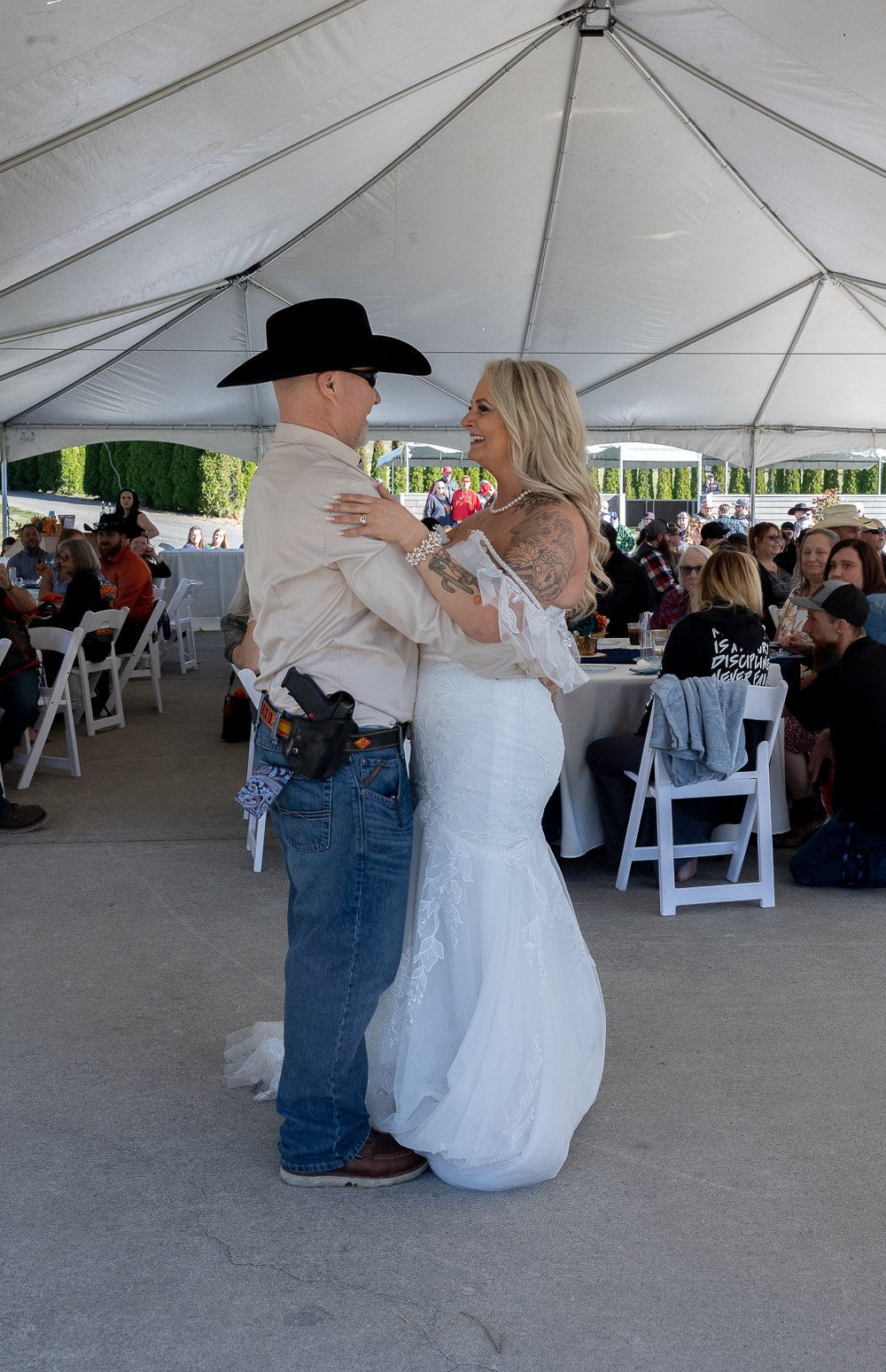 A couple dancing at their wedding reception inside a large tent with seated guests watching.