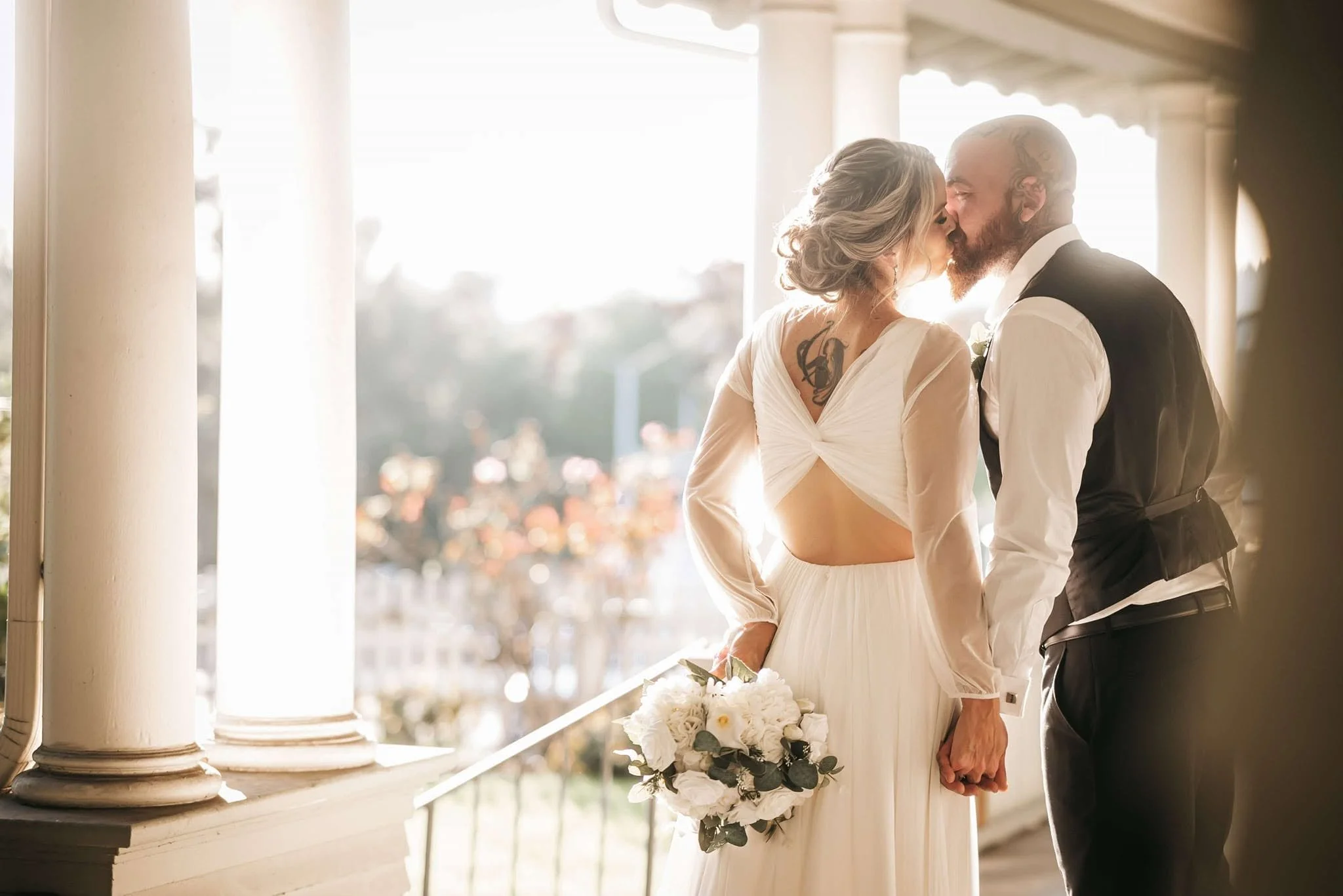 A bride and groom sharing a kiss on their wedding day, standing on a balcony with sunlight streaming through columns, with the bride holding a bouquet of white flowers.