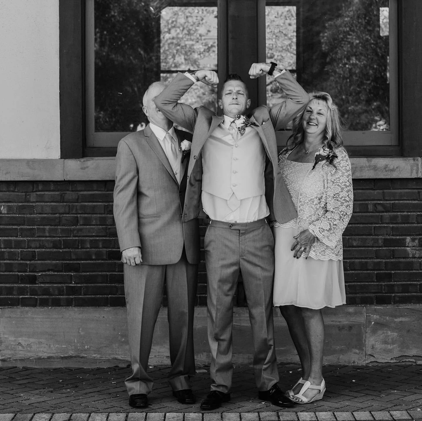 Black and white photo of an older man, a younger man, and a woman standing outside in front of a brick wall and a window. The younger man is flexing his arms confidently, while the older man and woman smile. The older man and woman are dressed in for