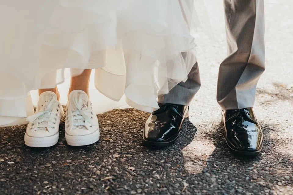 Close-up of the feet of a bride and groom standing on a gravel surface, with the bride wearing white sneakers and the groom wearing shiny black formal shoes.