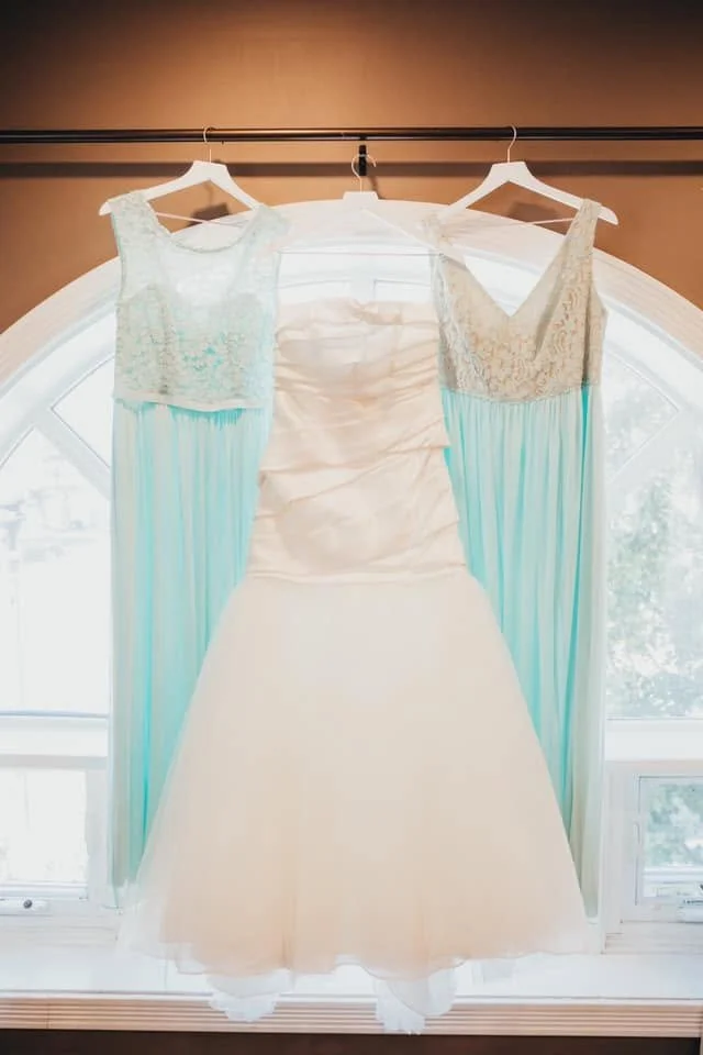 A white wedding dress hanging in front of a window, flanked by two blue and white ribbons or curtains.