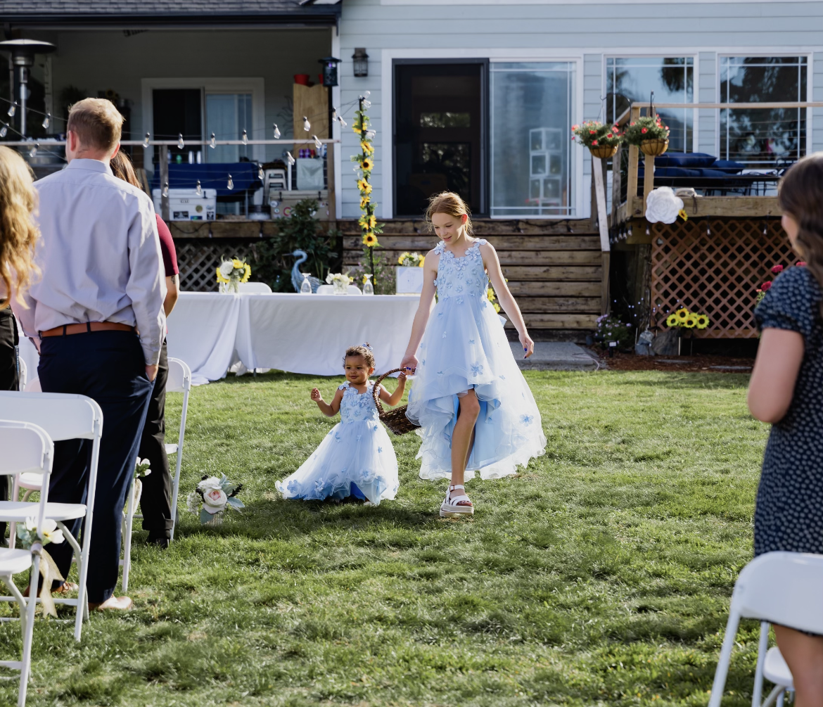 A young girl and a smaller girl walk in a garden wedding reception, holding hands. The girl is wearing a light blue dress with floral details and sandals, while the smaller girl wears a similar dress and shoes. Guests stand around them, and a table w