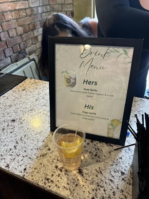 A drink menu sign on a counter with a glass of water with lemon in front of it, in a restaurant or bar setting.