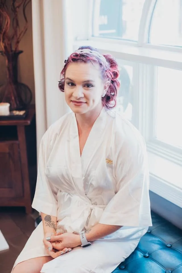 A woman with pink and purple curled hair sitting on a dark blue tufted bench near a window, wearing a white robe, with a tattoo on her left forearm.