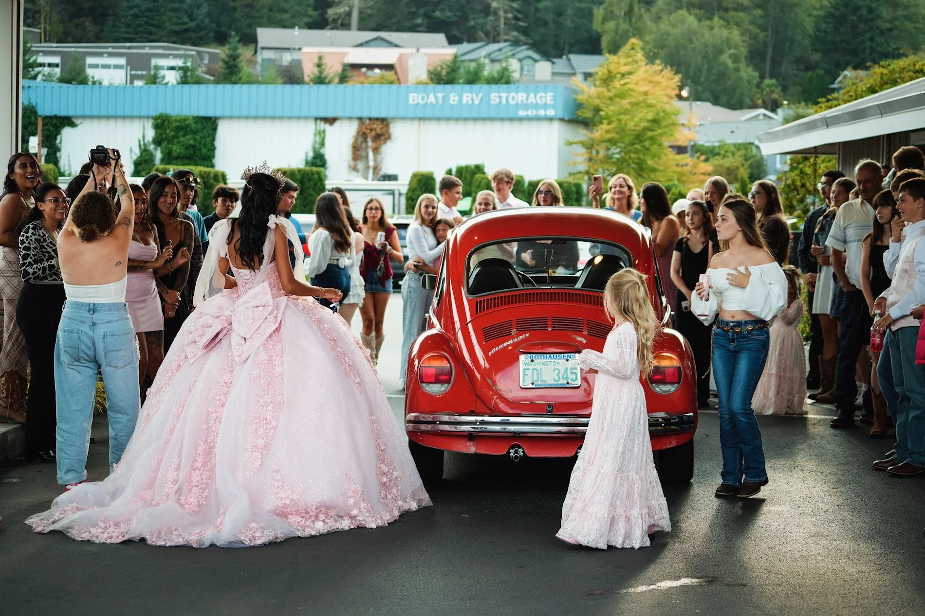 A wedding scene with a woman in a pink ball gown dress, a young girl in a white dress, and a vintage red Volkswagen Beetle surrounded by a crowd of onlookers, some taking photos. The scene appears to be a parade or public celebration outside a buildi