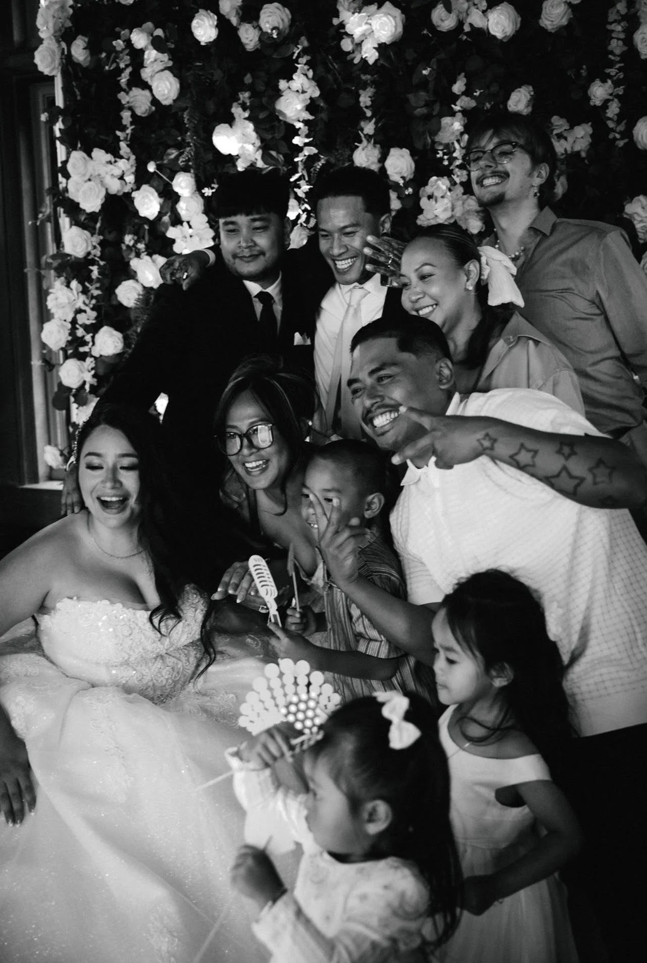 A joyful group of people celebrating a wedding, including a bride in a wedding dress, surrounded by friends and family, with a floral backdrop.