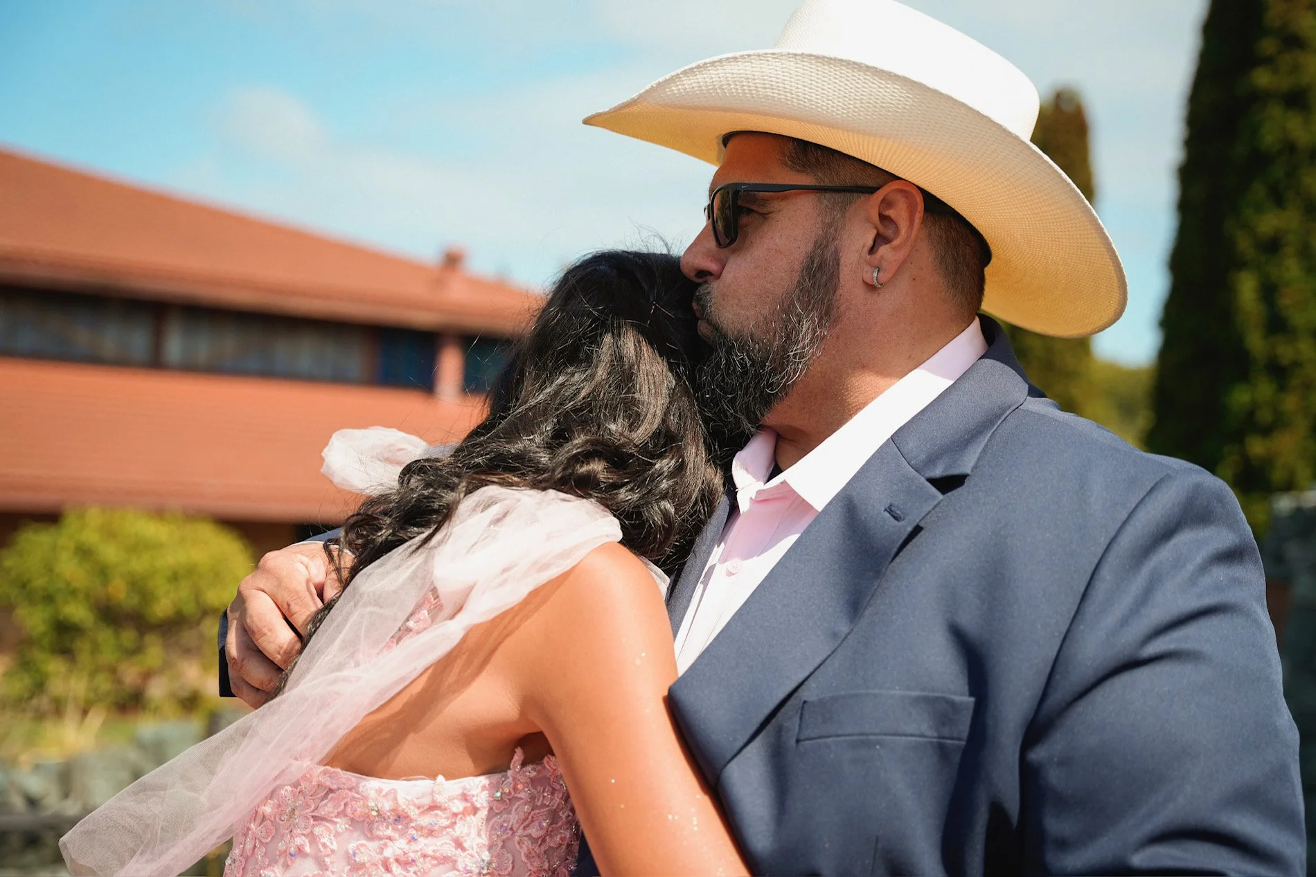 A man wearing a cowboy hat and sunglasses is hugging a woman in a pink dress with lace and embroidery. They are outdoors under a clear sky, with a building with a red roof and green trees in the background.