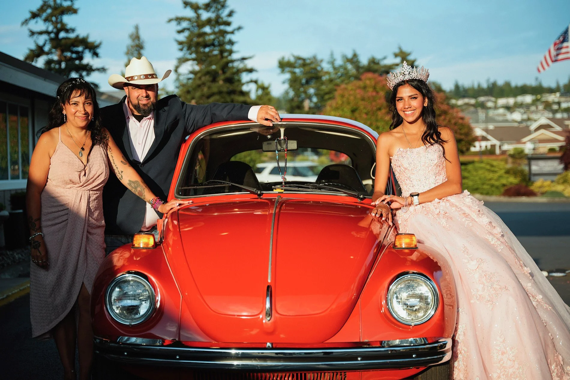 Group of four people posing with a vintage red car outdoors. One woman in a pink dress and young woman in a wedding gown and tiara on the right. Two adults, a man with a cowboy hat and a woman in a pink dress, on the left. Background with trees, hous