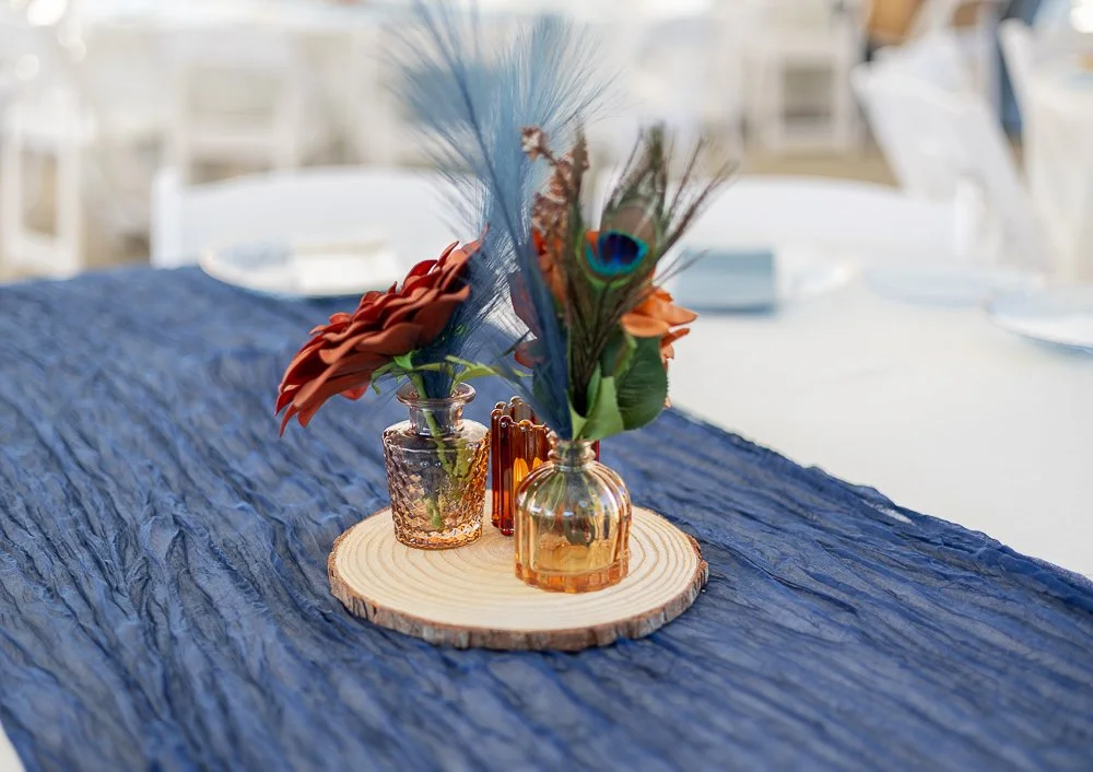 Table centerpiece with flowers and peacock feather in amber and pink glass vases on a wooden slab, set on a blue textured table runner.