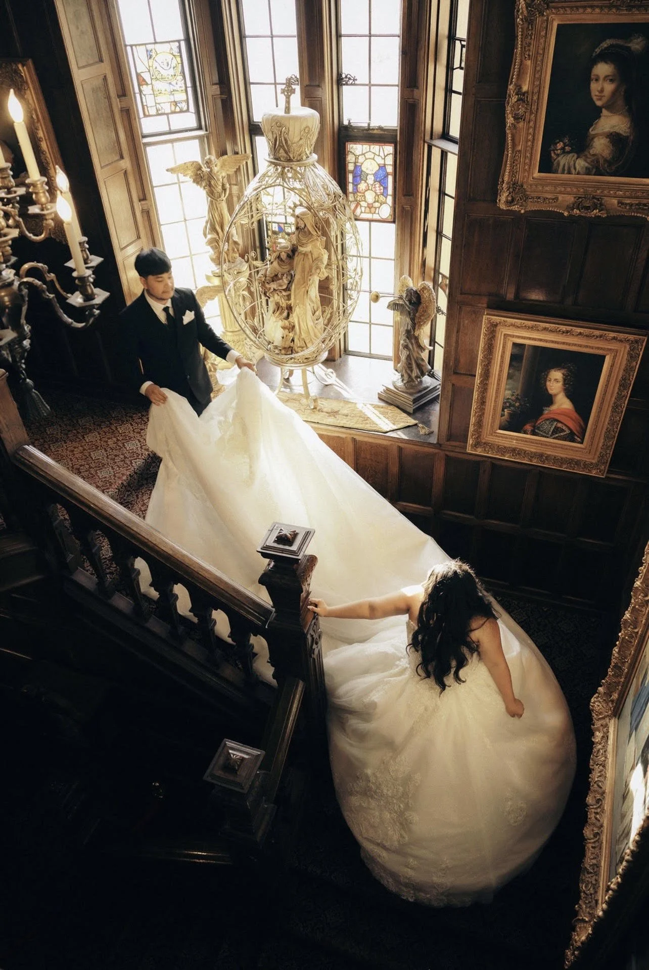 A bride in a wedding gown walking up a staircase while a man in a tuxedo stands at the top holding the train of her dress. The staircase is decorated with ornate wood paneling and framed portraits. Large stained-glass windows let in natural light, an