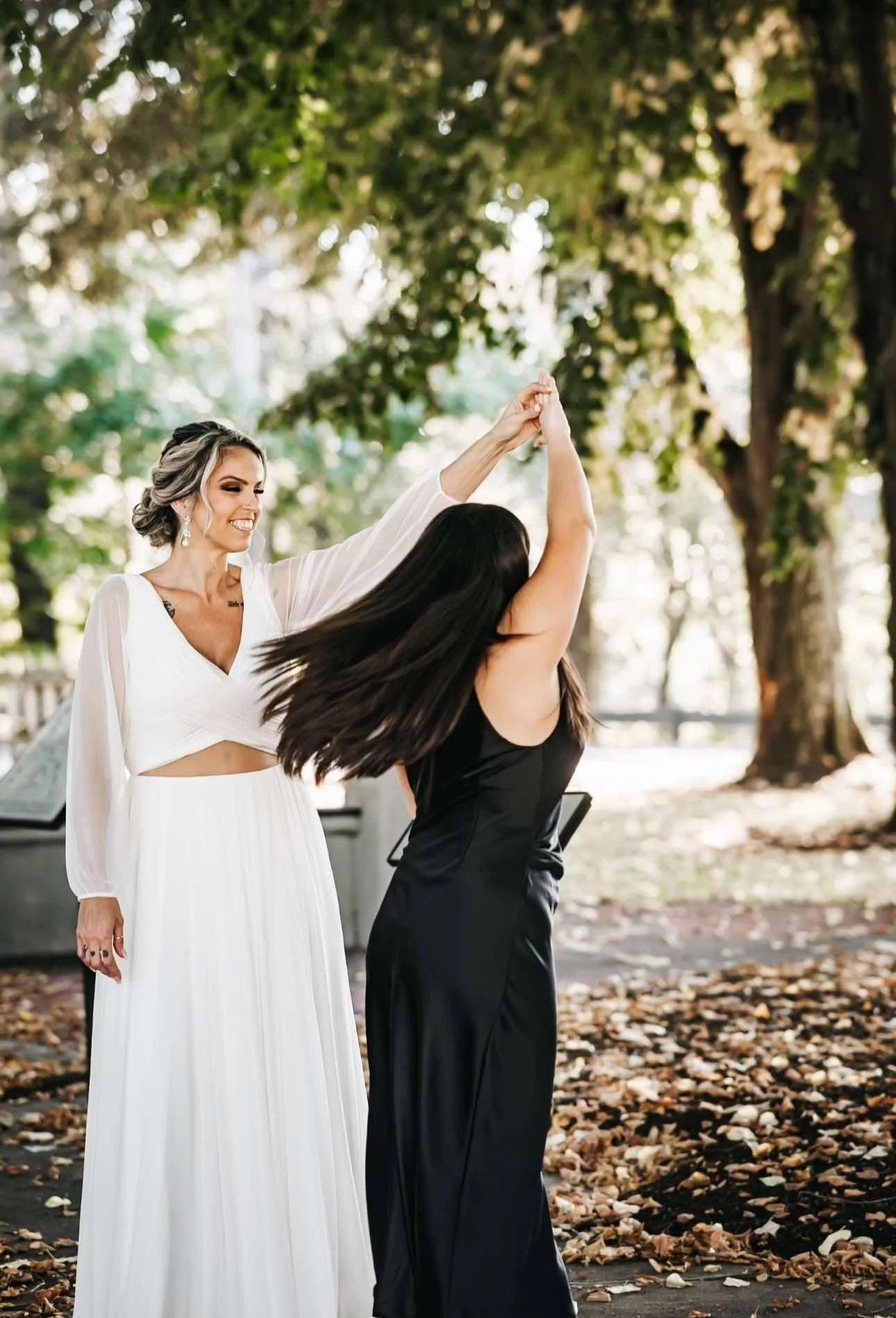 A woman in a white dress dancing with a woman in a black dress outdoors in a park with trees and fallen leaves.