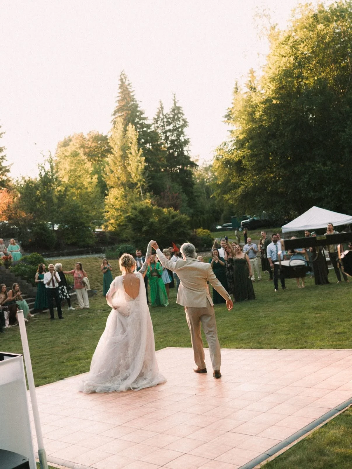 A bride and groom dancing at an outdoor wedding reception with guests watching in the background, surrounded by trees and a white tent.