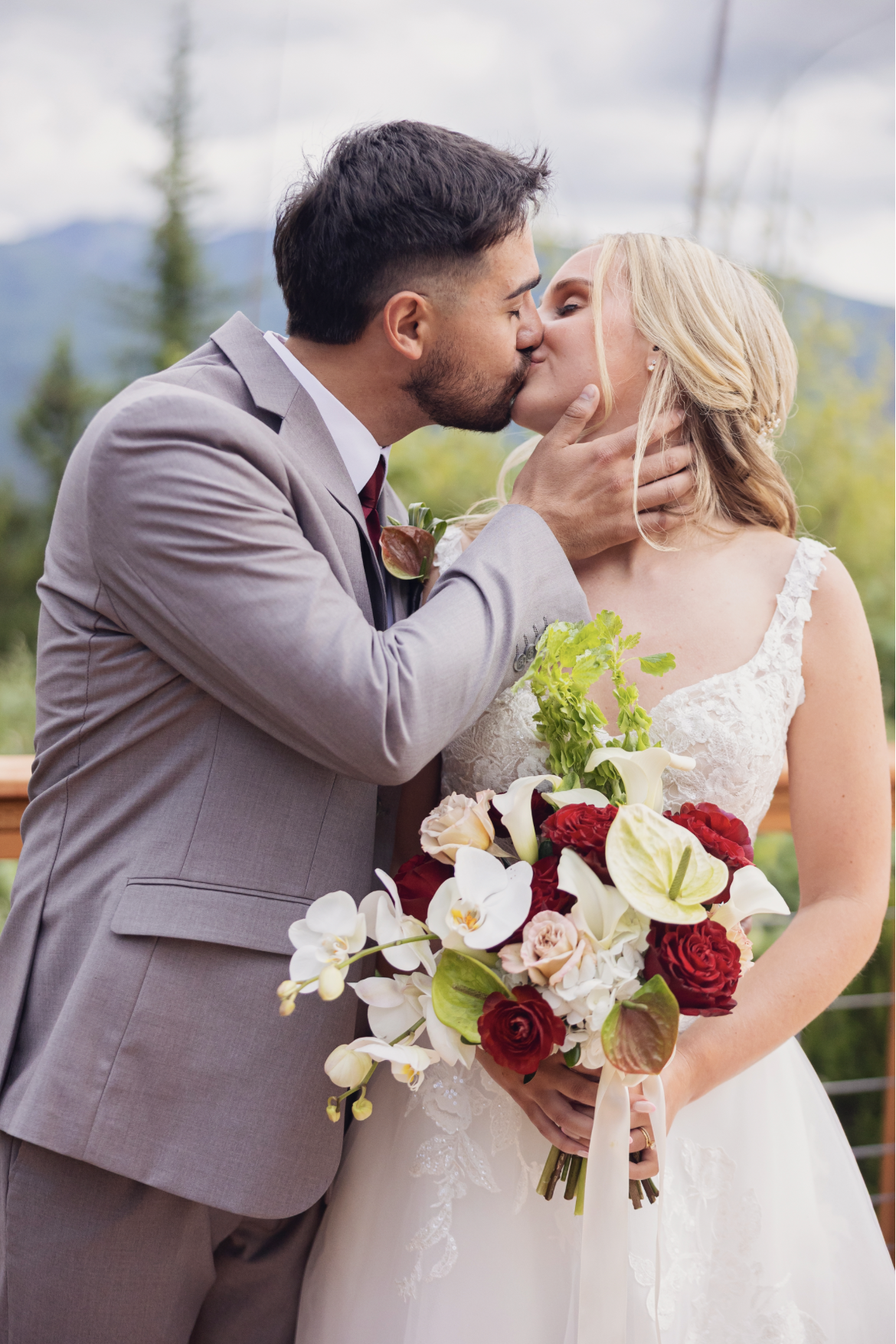 A newlywed couple sharing a kiss outdoors, with the groom holding the bride's face and wearing a gray suit, while the bride, dressed in a white gown, holds a large bouquet of flowers including red, white, and pink roses, white calla lilies, and greenery, with lush trees and mountains in the background.
