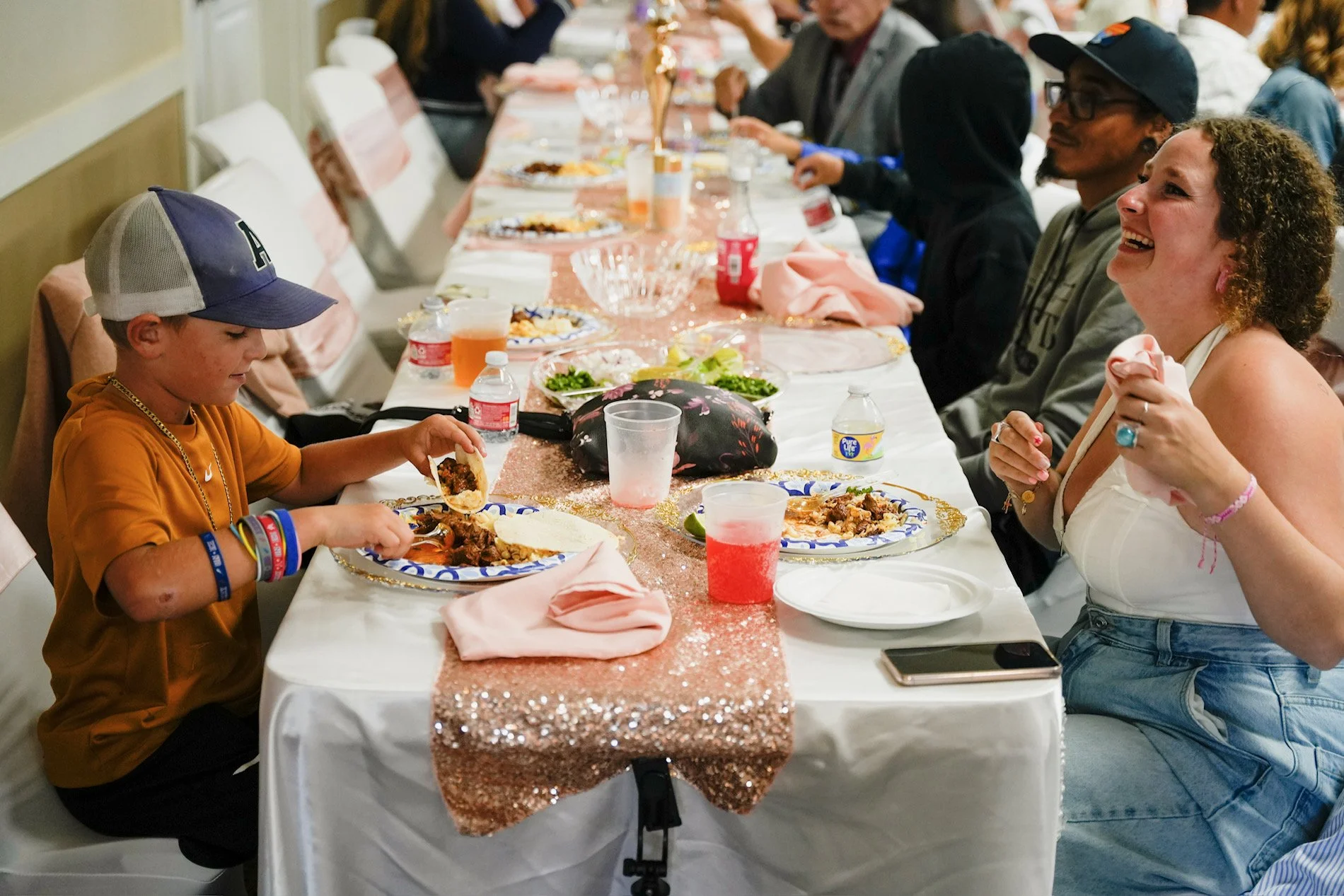 People sitting at a long table enjoying a meal at a celebration or gathering, with food, drinks, and smiles.