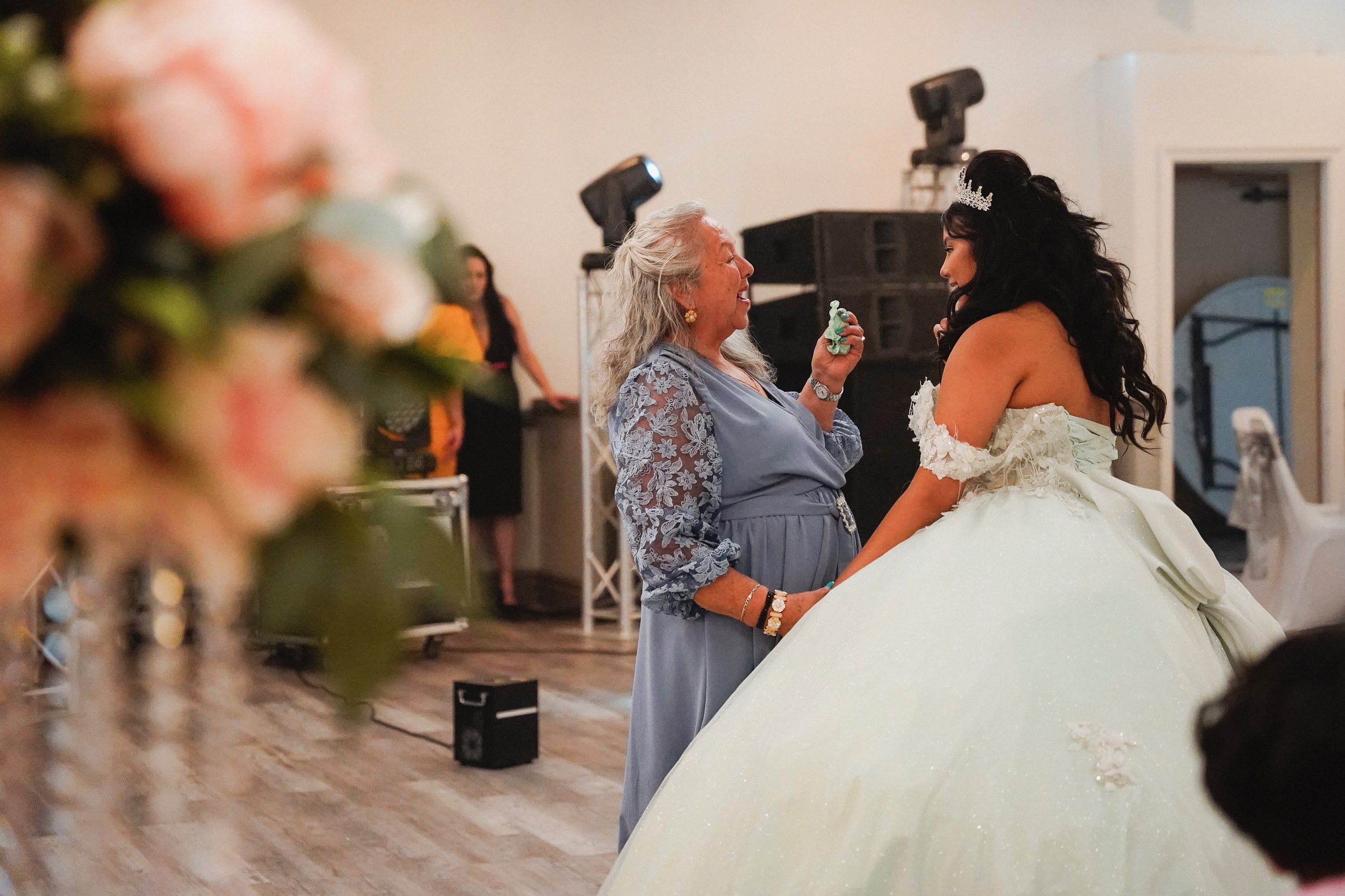 An elderly woman and a young woman, dressed as a bride, smiling and holding hands at a wedding reception, with speakers and a partially visible person in the background.