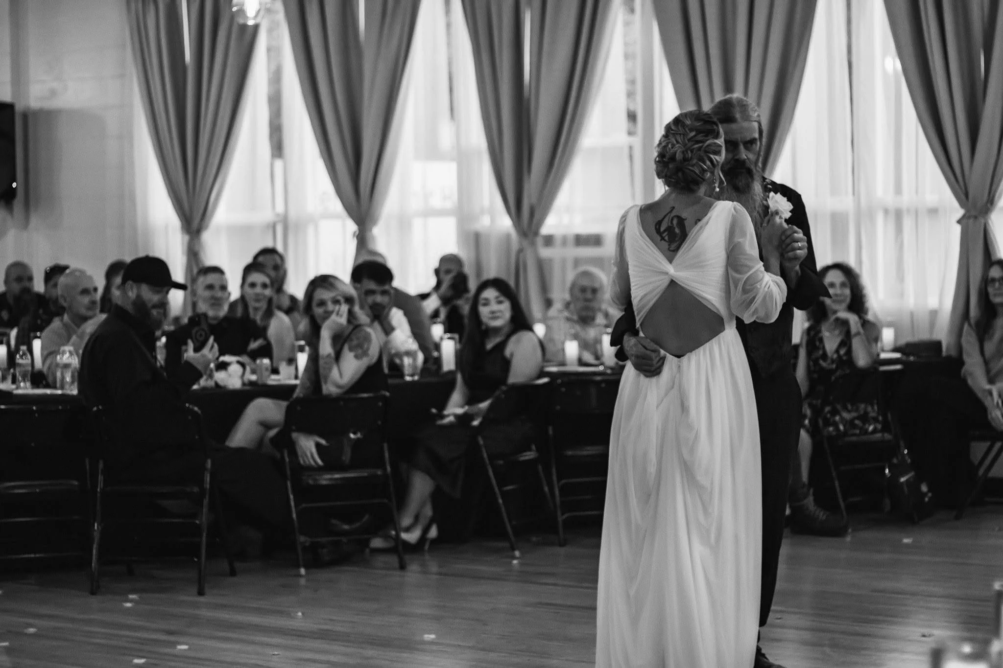 A bride and groom dance together in a large, well-lit room with tall curtains and many guests seated at tables, watching them.
