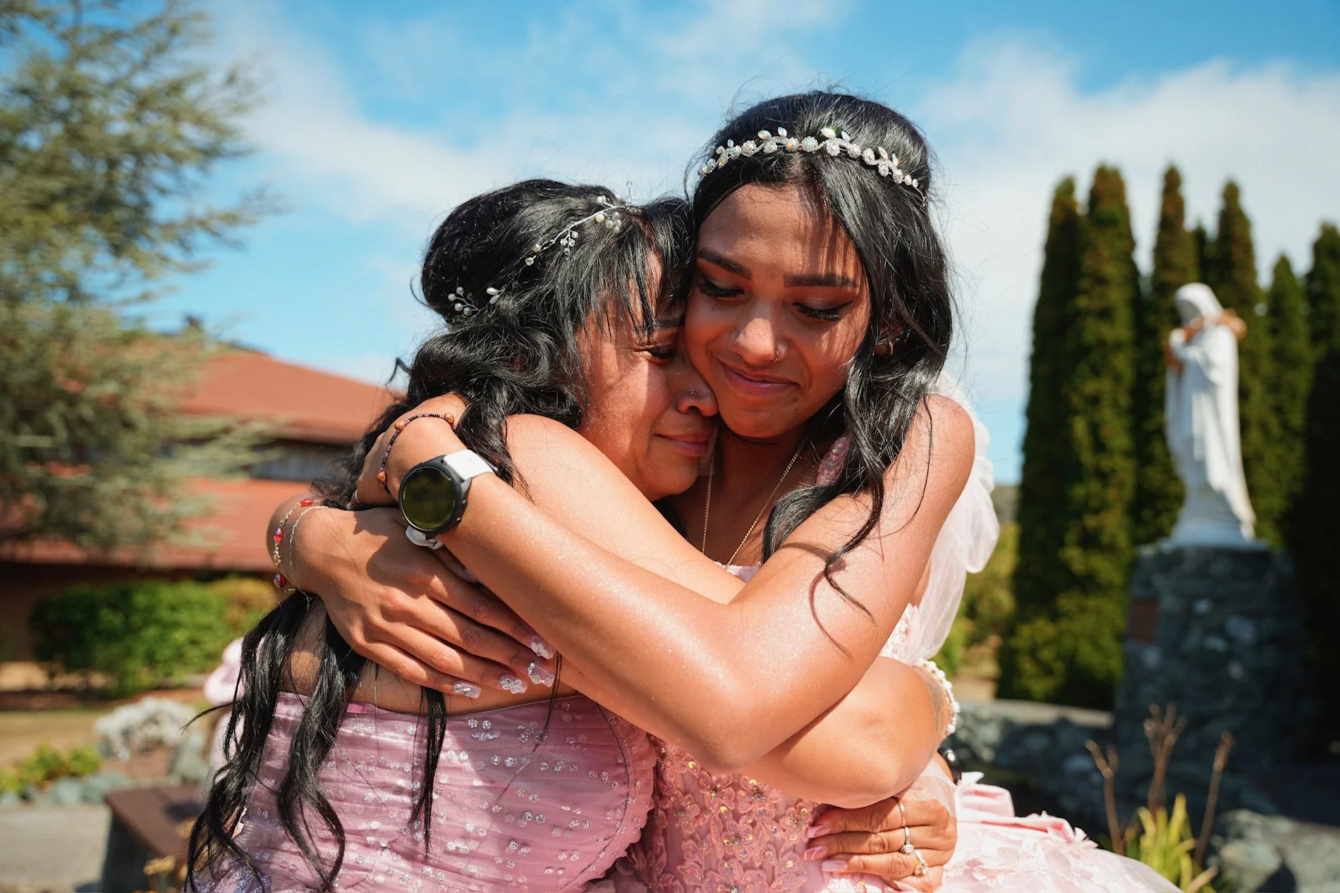Two women with long black hair wearing floral headbands, hugging each other outdoors under a blue sky, with a statue and trees in the background.