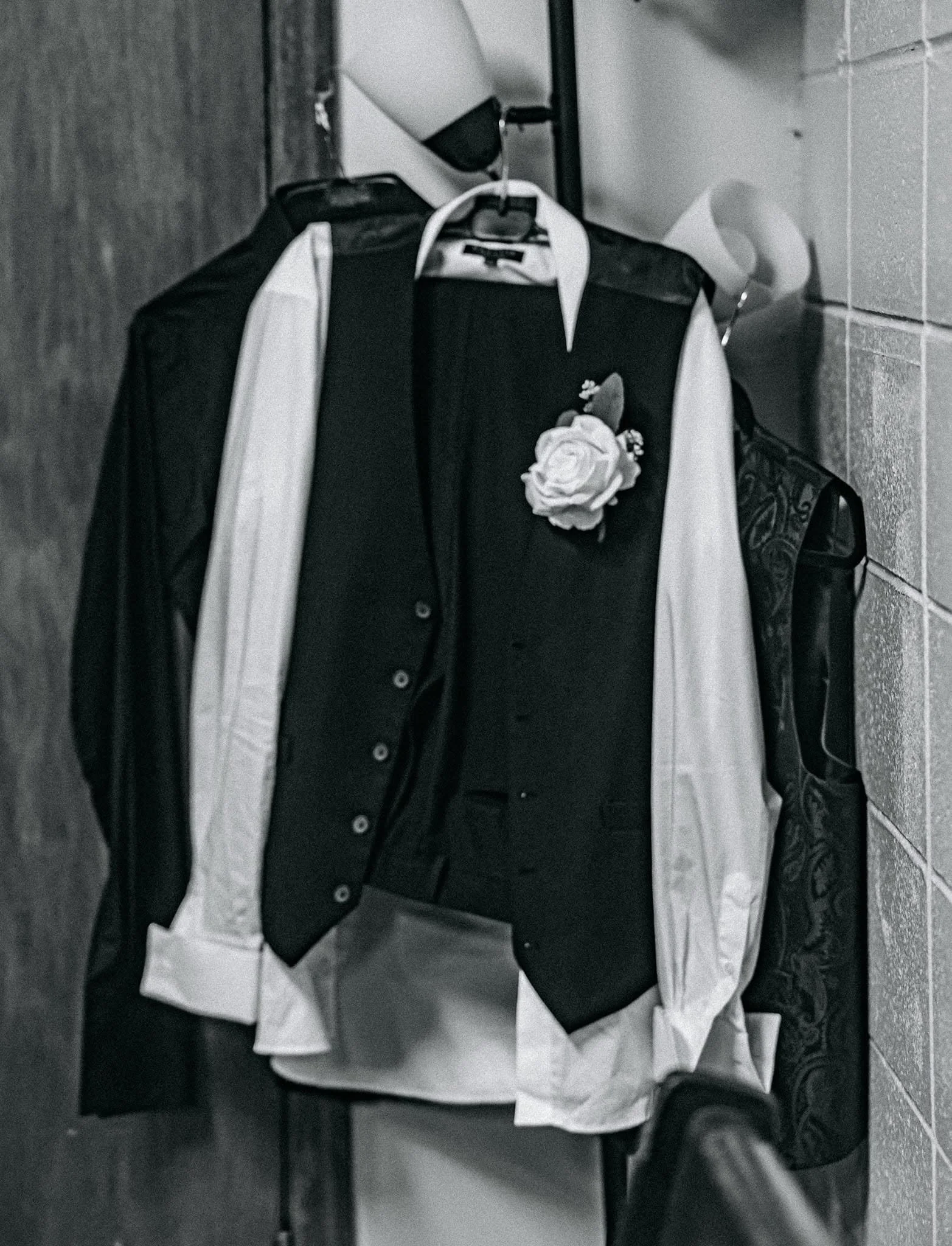 Black and white photo of a clothing rack with a jacket with a white flower brooch on it, a white shirt, and other garments hanging.