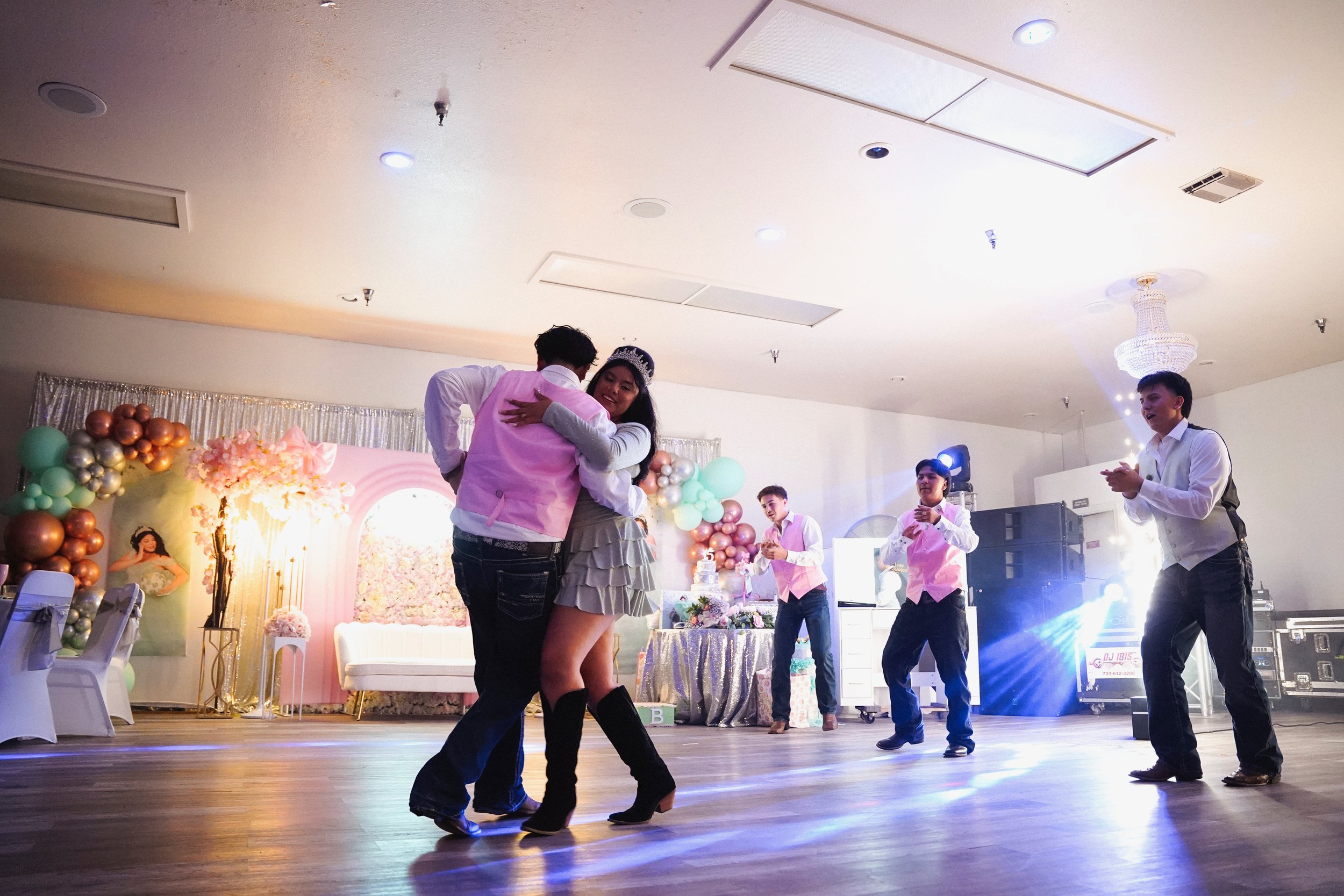 People dancing and celebrating at a decorated event with balloons, flowers, and a stage, with a woman wearing a tiara and a man in a pink vest.