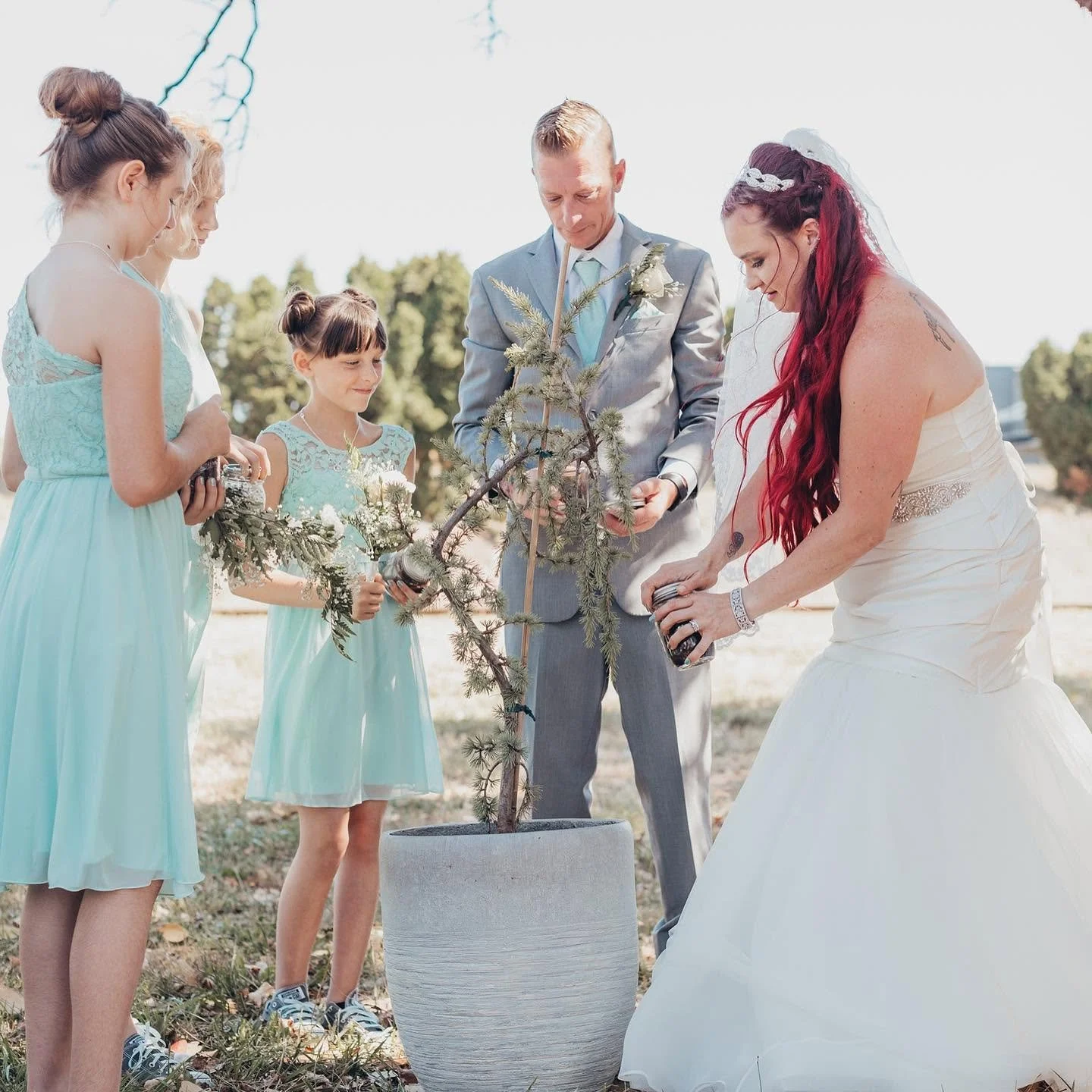 A wedding ceremony outdoors with a bride, groom, and three young bridesmaids planting a small tree together.