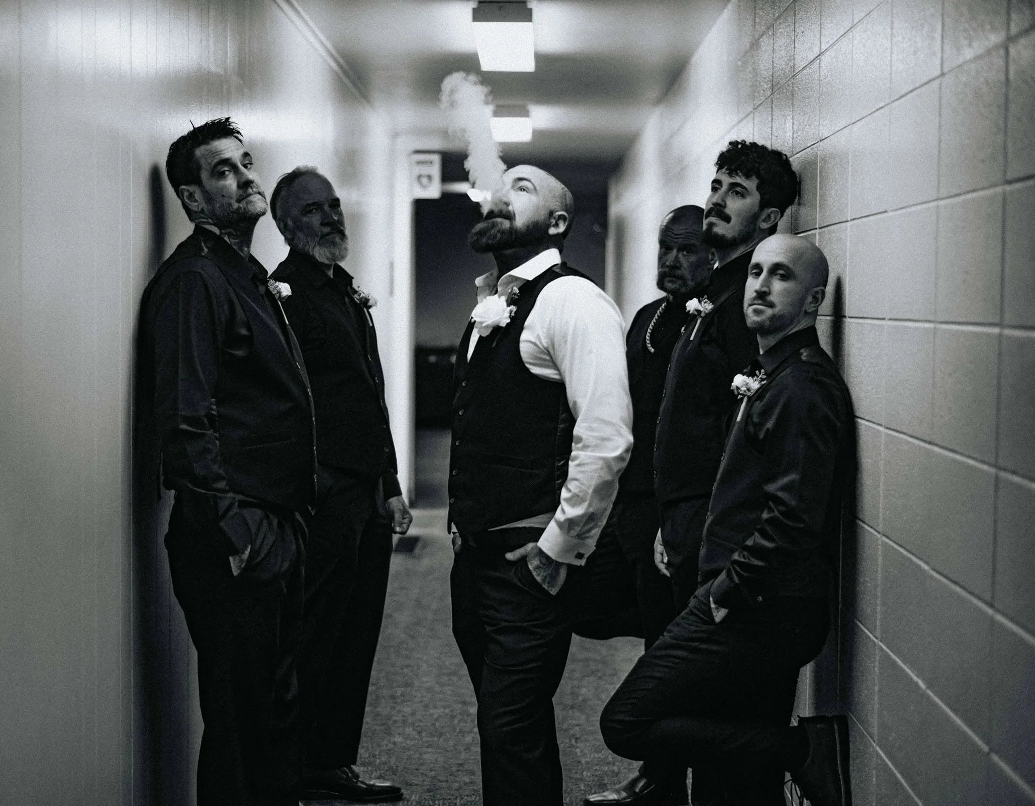 Five men wearing black clothing and corsages standing in a line against tiled walls in a dimly lit hallway, with one man in the center smoking a cigarette.