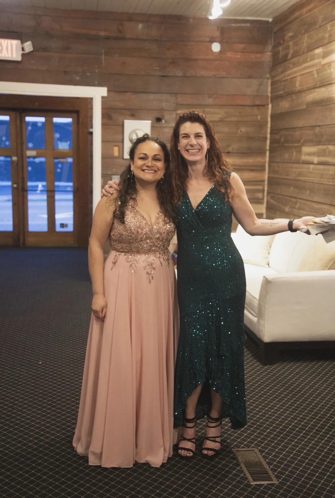 Two women in elegant evening dresses standing inside a room with wooden walls, smiling at the camera. One woman is wearing a light pink gown with lace detail and the other is wearing a dark green, sparkly gown. They are standing close together with a