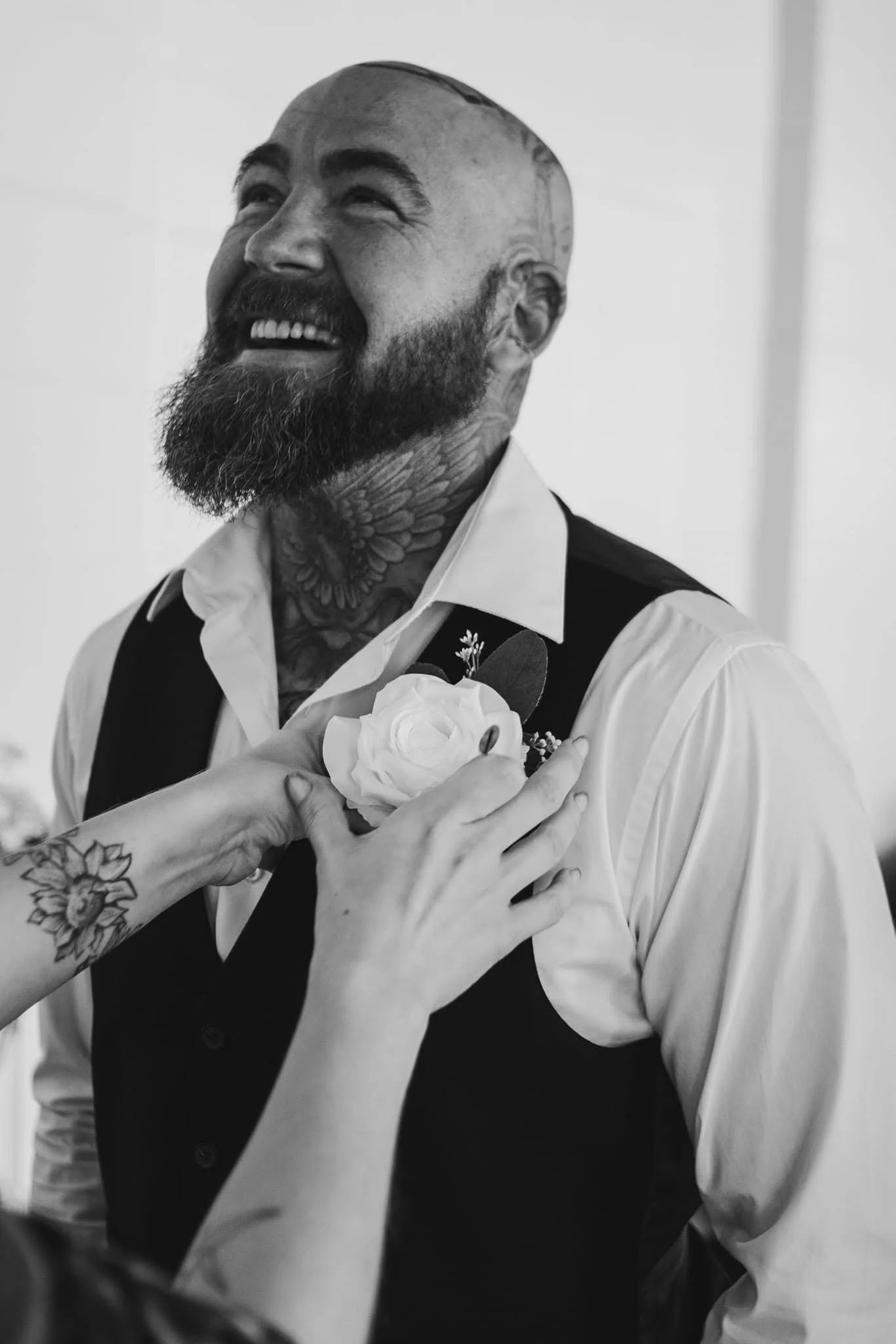 Black-and-white photo of a smiling man with tattoos, a beard, and a shaved head, holding a flower while someone else's hand touches his chest.