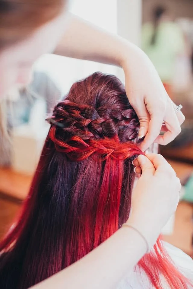 A hairstylist braiding a woman's long, red-colored hair.