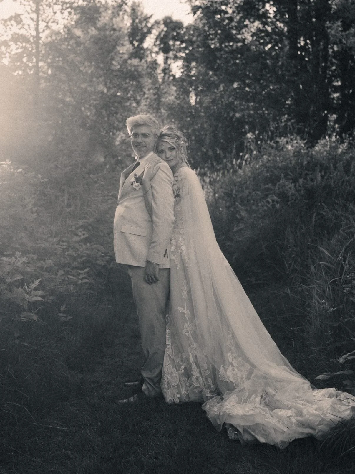 A black and white photo of a bride and groom standing outdoors surrounded by trees and foliage. The bride is leaning against the groom's shoulder, both are dressed in wedding attire, with the bride in a long lace gown and the groom in a light-colored