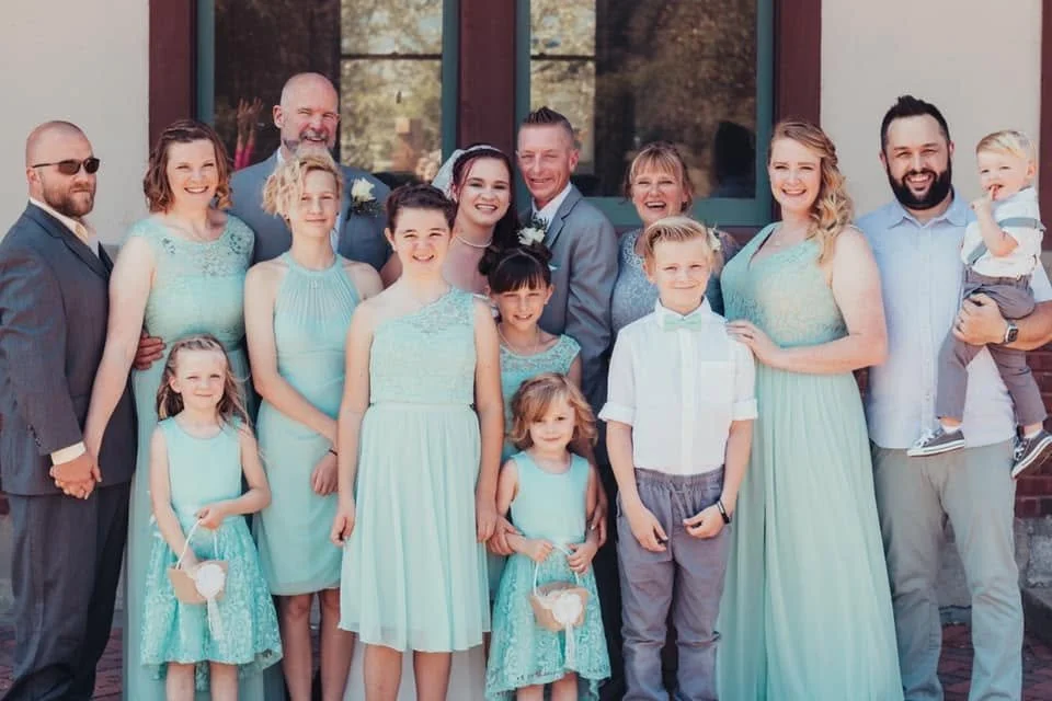 Group of people, including children and adults, dressed in pastel and formal attire, standing outside a building with large windows, smiling for a photo.