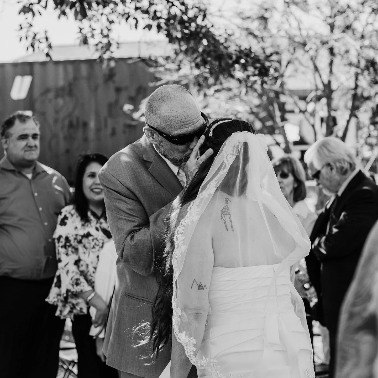 A couple shares a kiss during their wedding ceremony, with guests watching and smiling in the background, outdoors with trees visible.
