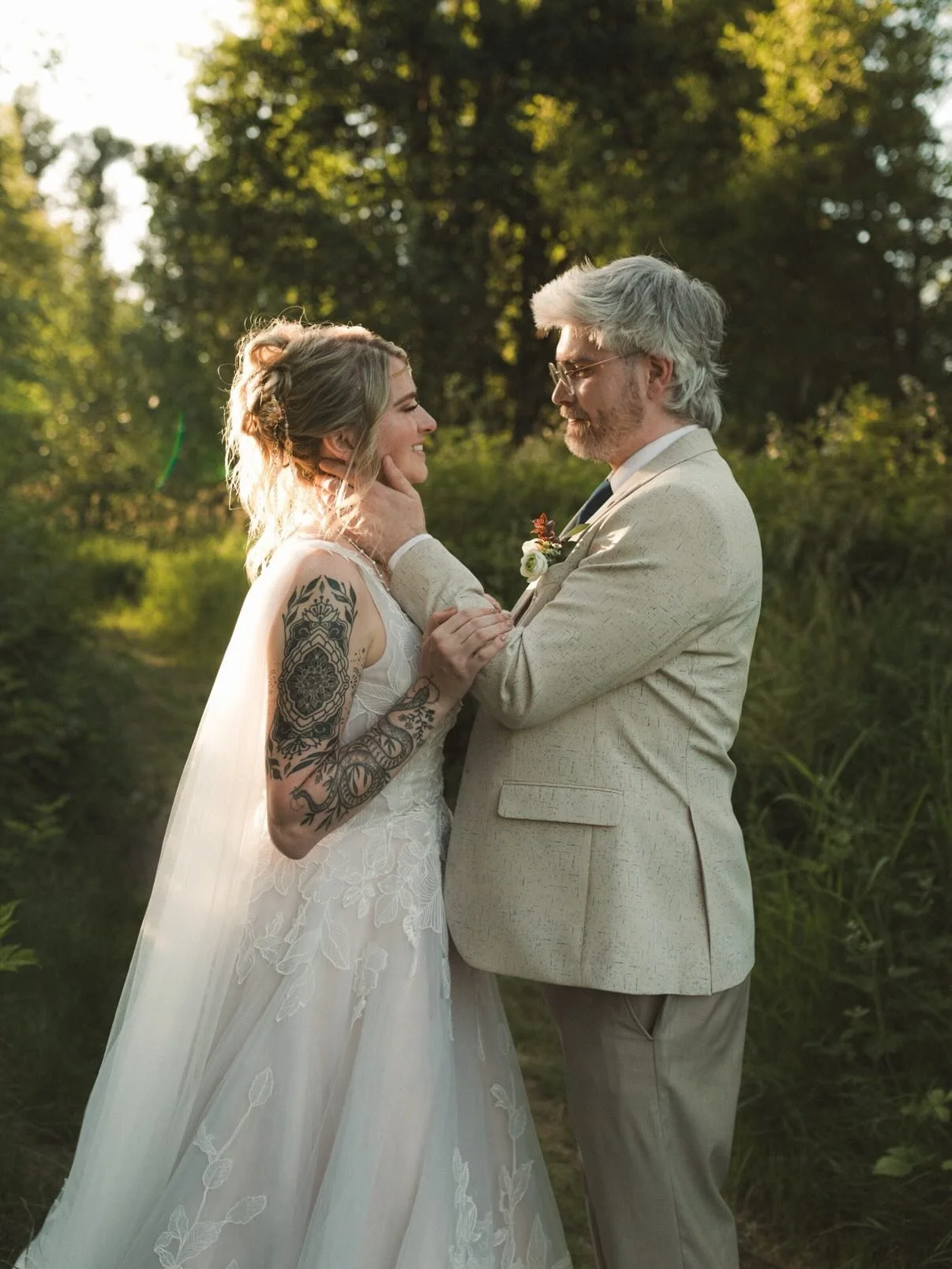 A bride and groom sharing an intimate moment outdoors during sunset, with the bride smiling and the groom holding her face gently, surrounded by greenery.