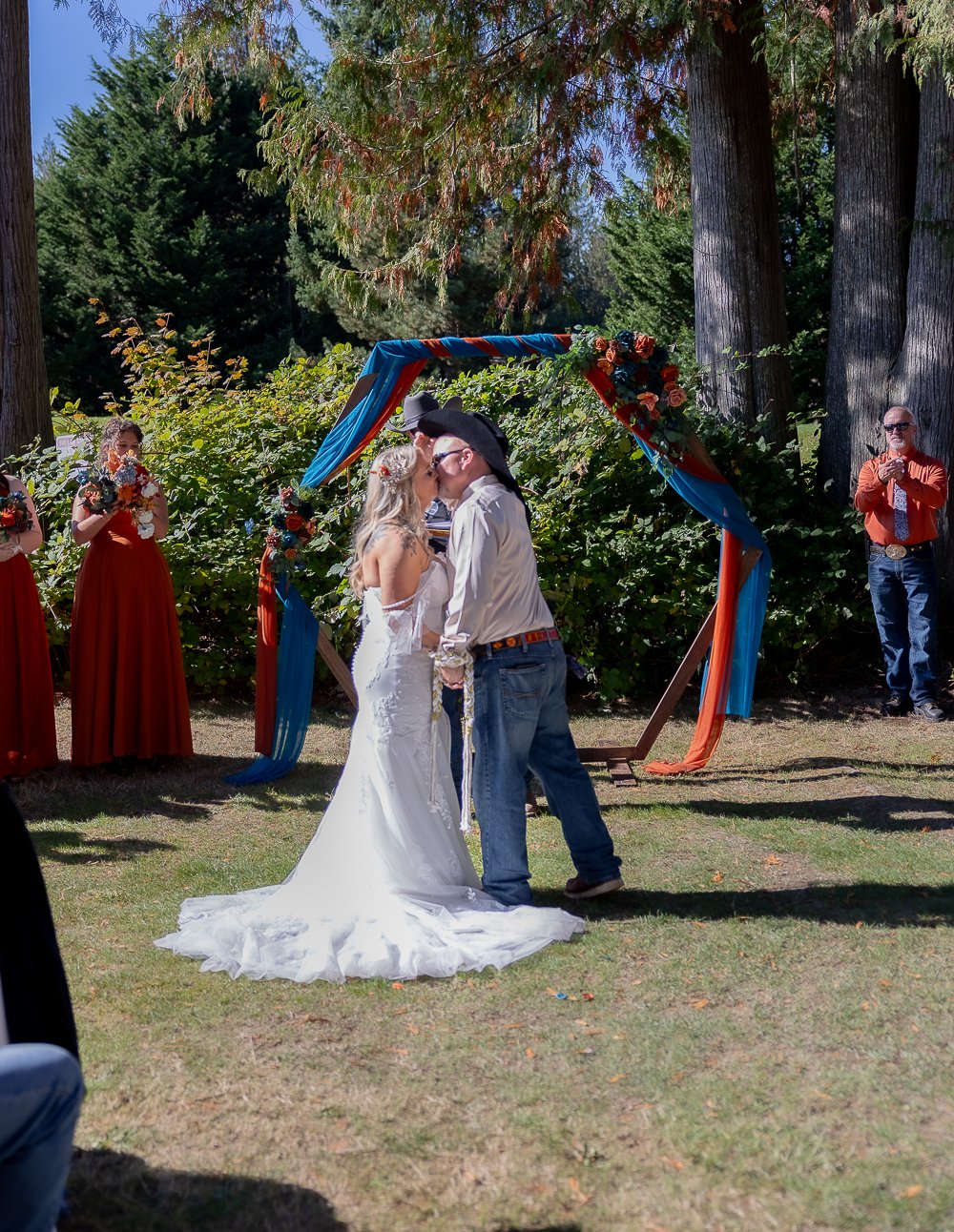A couple dressed in wedding attire share a kiss during an outdoor wedding ceremony, with bridesmaids in red dresses holding bouquets and guests taking photos, set amidst trees and floral decorations.