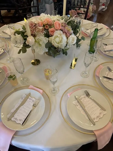 Round table set for a formal event with white tablecloth, floral centerpiece with pink and white flowers, candles, and place settings including plates, silverware, glasses, and pink napkins.