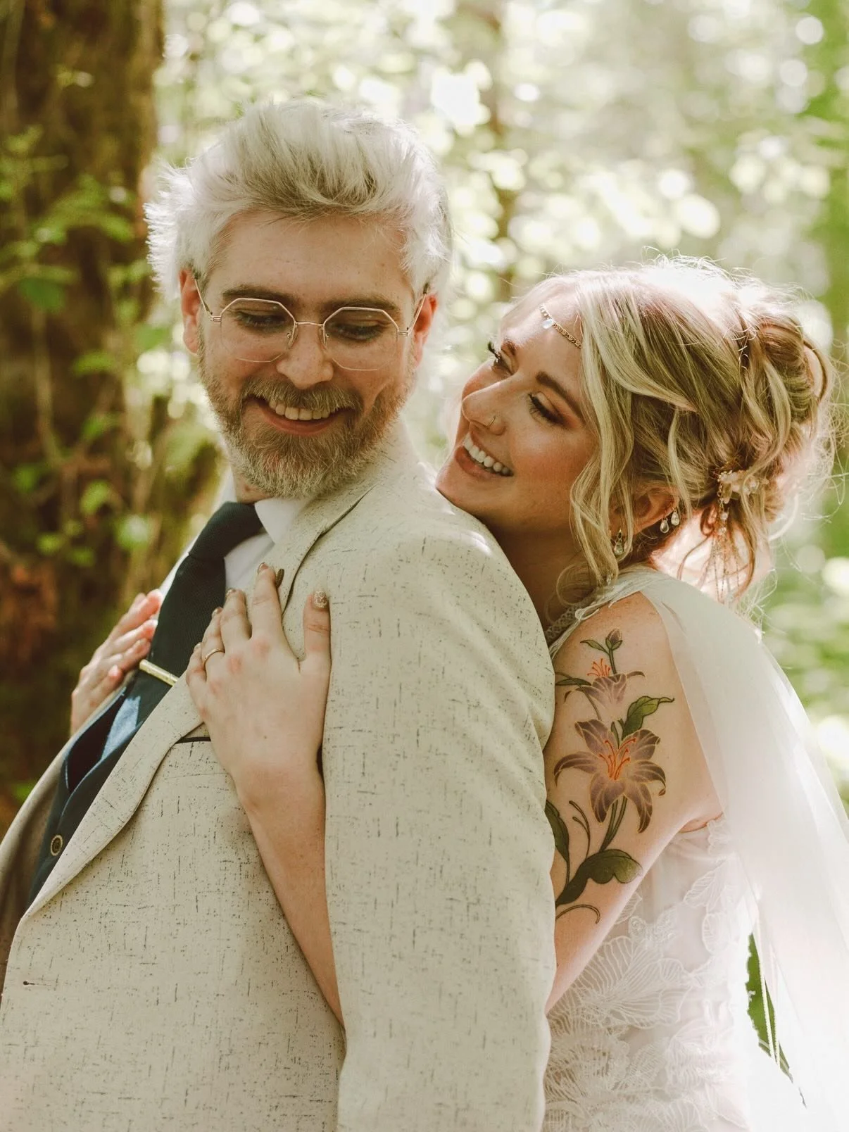 A happy couple, possibly newlyweds, embracing outdoors in a forest setting, with the woman smiling at the man and showing a floral tattoo on her arm.