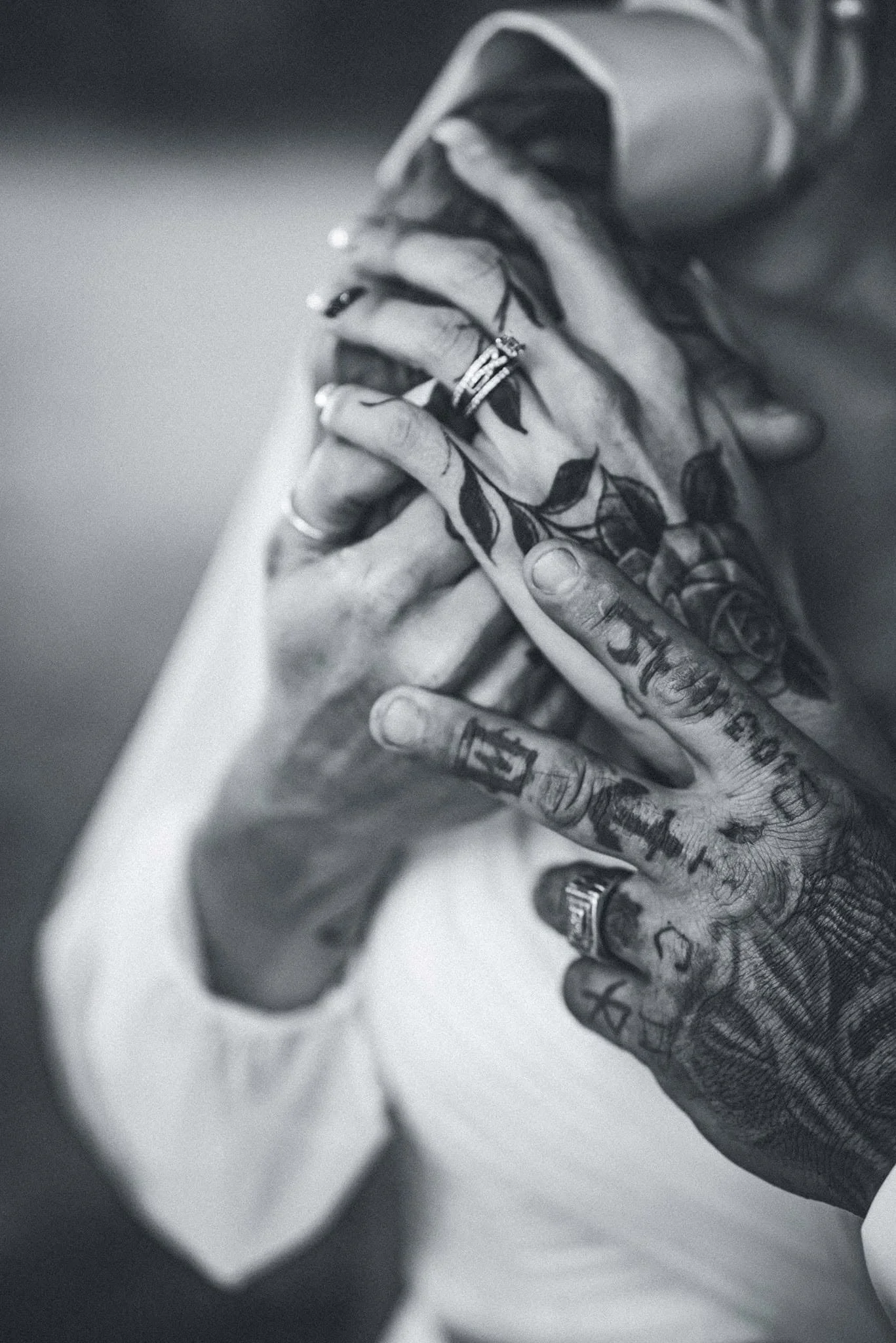 Close-up of tattooed hands holding a smartphone, with rings on fingers and visible tattoos on hands and wrists, in black and white.
