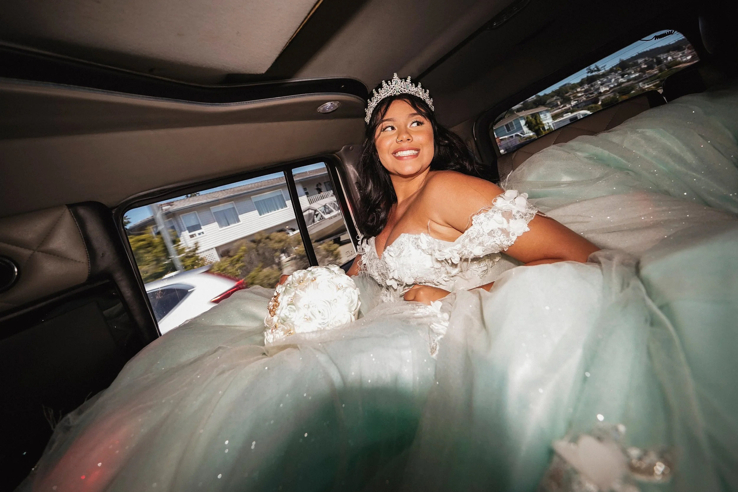 A woman wearing a wedding dress and a tiara sitting inside a vehicle, smiling and looking out the window.