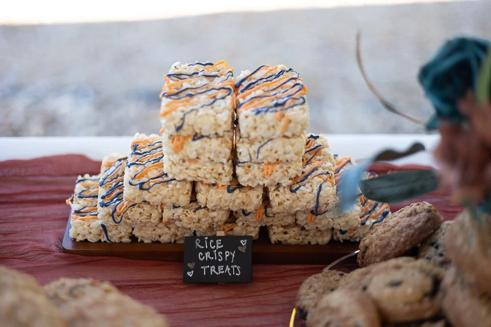 Stacks of rice crispy treats decorated with drizzles of chocolate and orange icing, displayed on a wooden board with a small sign reading 'Rice Crispy Treats' at an outdoor event.