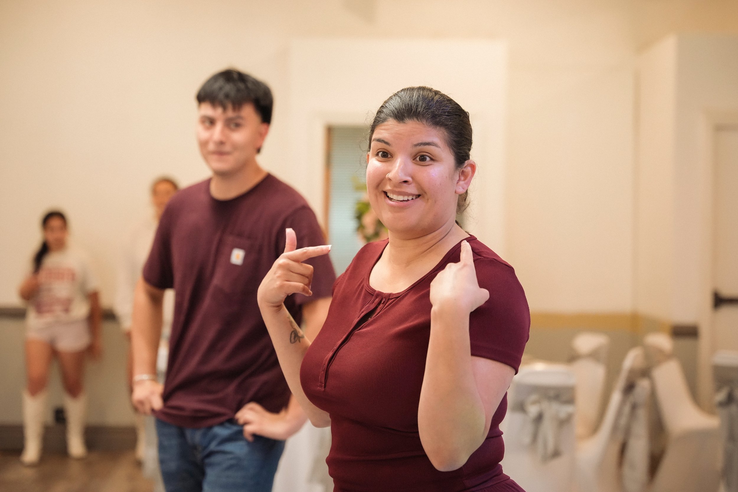 Two people in matching burgundy shirts standing indoors with blurred background. Woman in foreground smiling and pointing to her shoulders, man beside her looking towards camera.