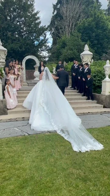 A wedding ceremony outdoors with a bride and groom at the center, surrounded by children and groomsmen, with a lush green background and a stone staircase.