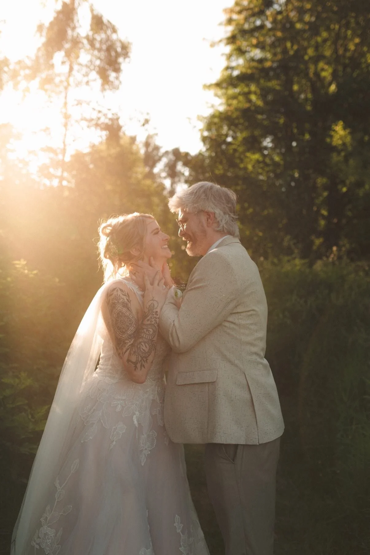 A bride and groom share an intimate moment outdoors at sunset, smiling at each other lovingly, with trees in the background and sunlight illuminating their faces.