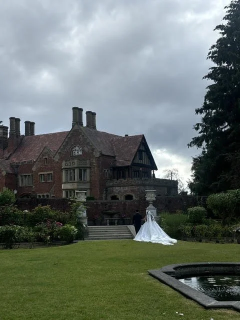 A large historic brick mansion with multiple chimneys and a gabled roof, surrounded by a well-maintained garden with a wedding dress displayed on a mannequin in front.